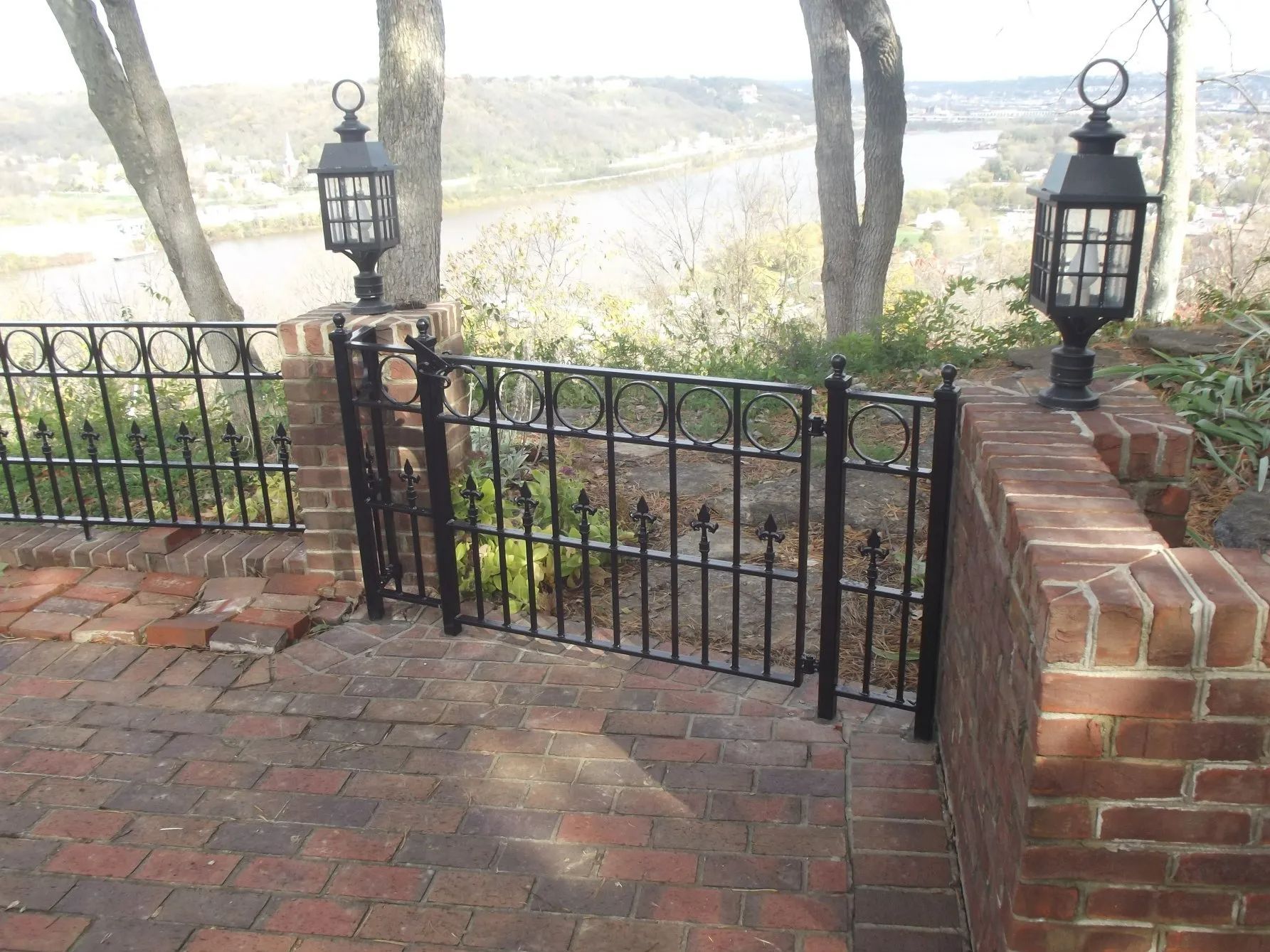 Black metal fence with brick pillars and lantern lights on a brick patio overlooking a scenic view.
