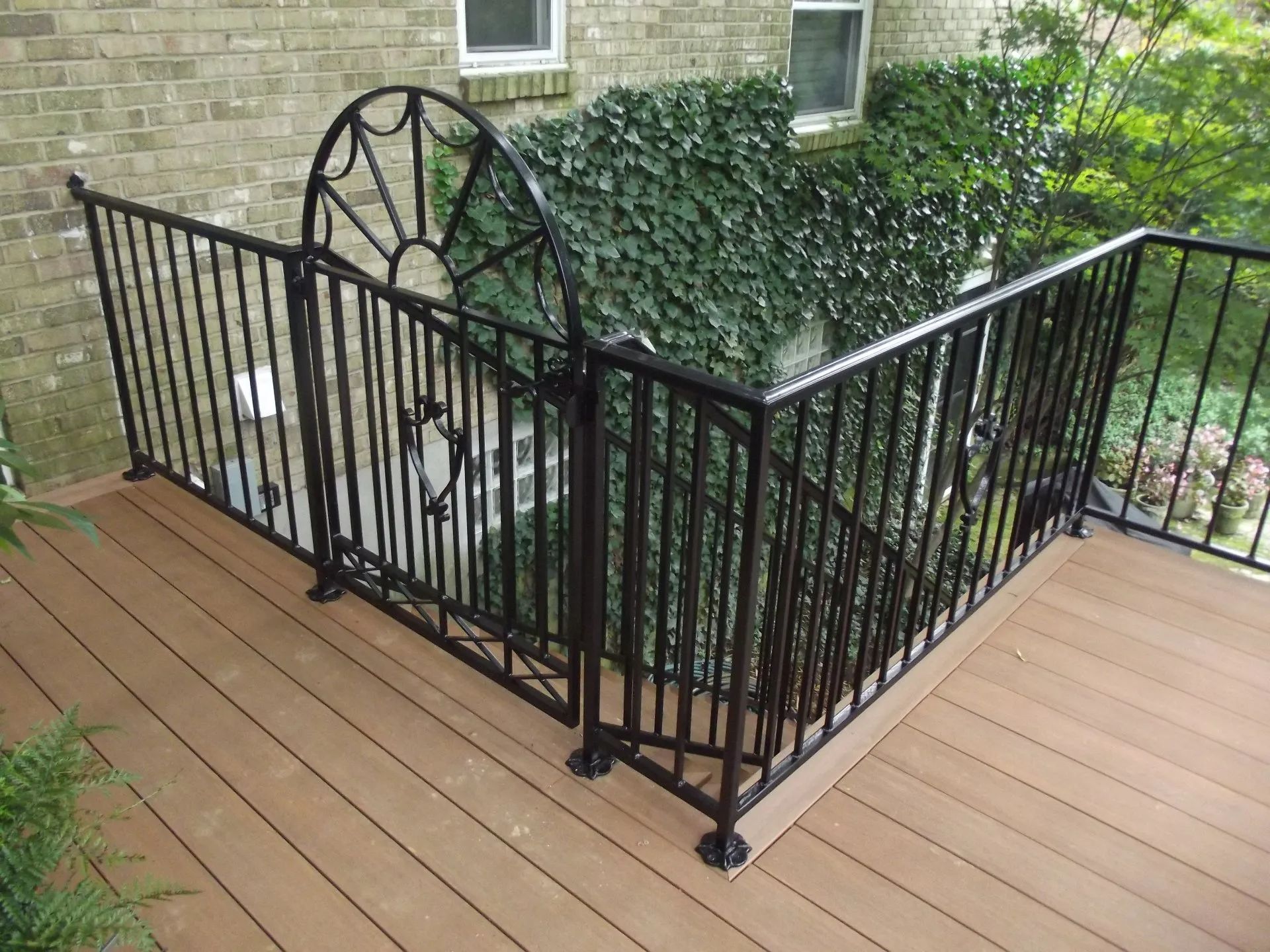 Black metal railing on a brown composite deck, with green ivy-covered brick wall backdrop.