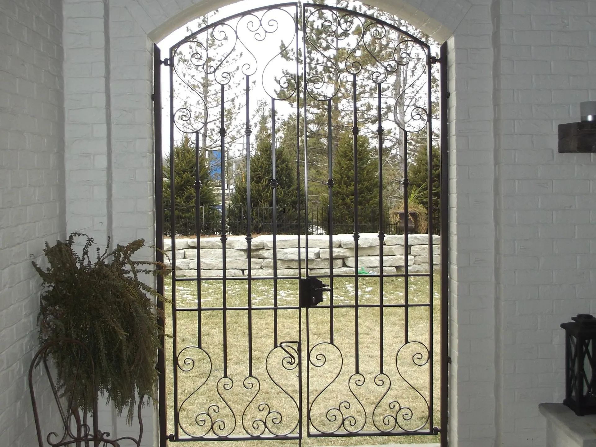 Black wrought iron gate set in a white brick archway, overlooking a yard with trees.