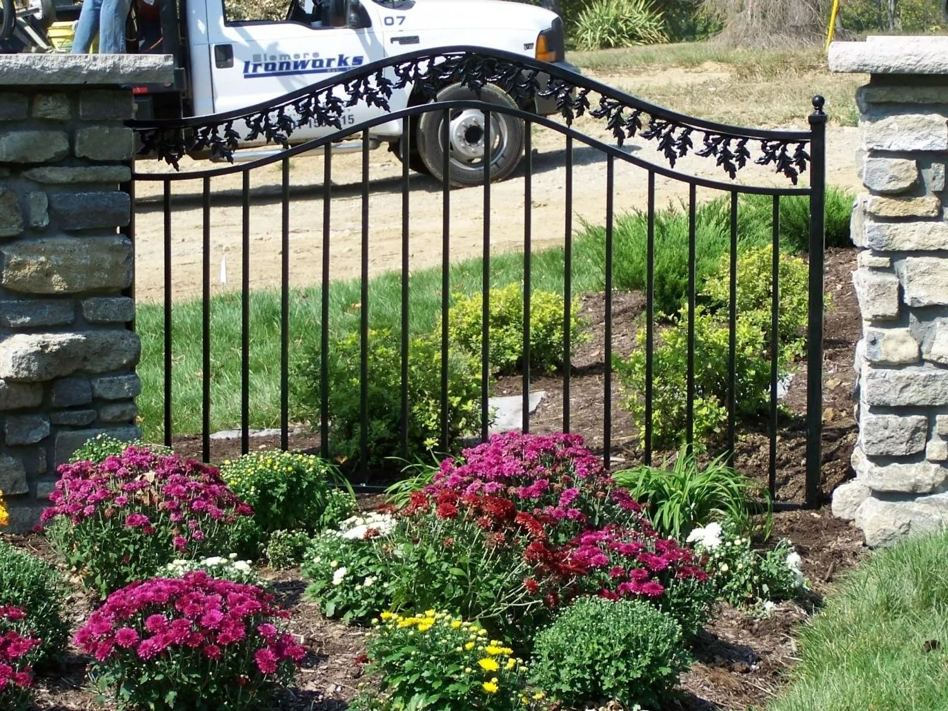 Black wrought iron gate with floral design, surrounded by stone pillars, a garden with purple and green flowers, and a white truck.