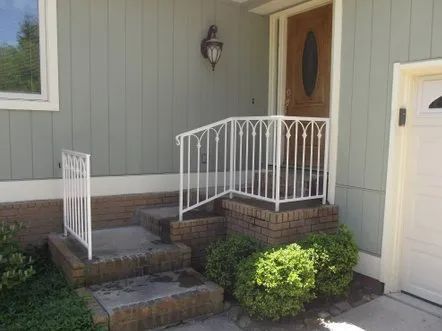 Exterior view of a house with white railings, brick steps, and a brown door. Green siding and bushes.