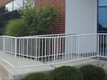 White metal railing around a small concrete ramp leading to a building's entrance; bushes in the foreground.