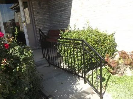 Black metal handrails on a concrete ramp leading to a brick building's entrance. Green bushes surround.