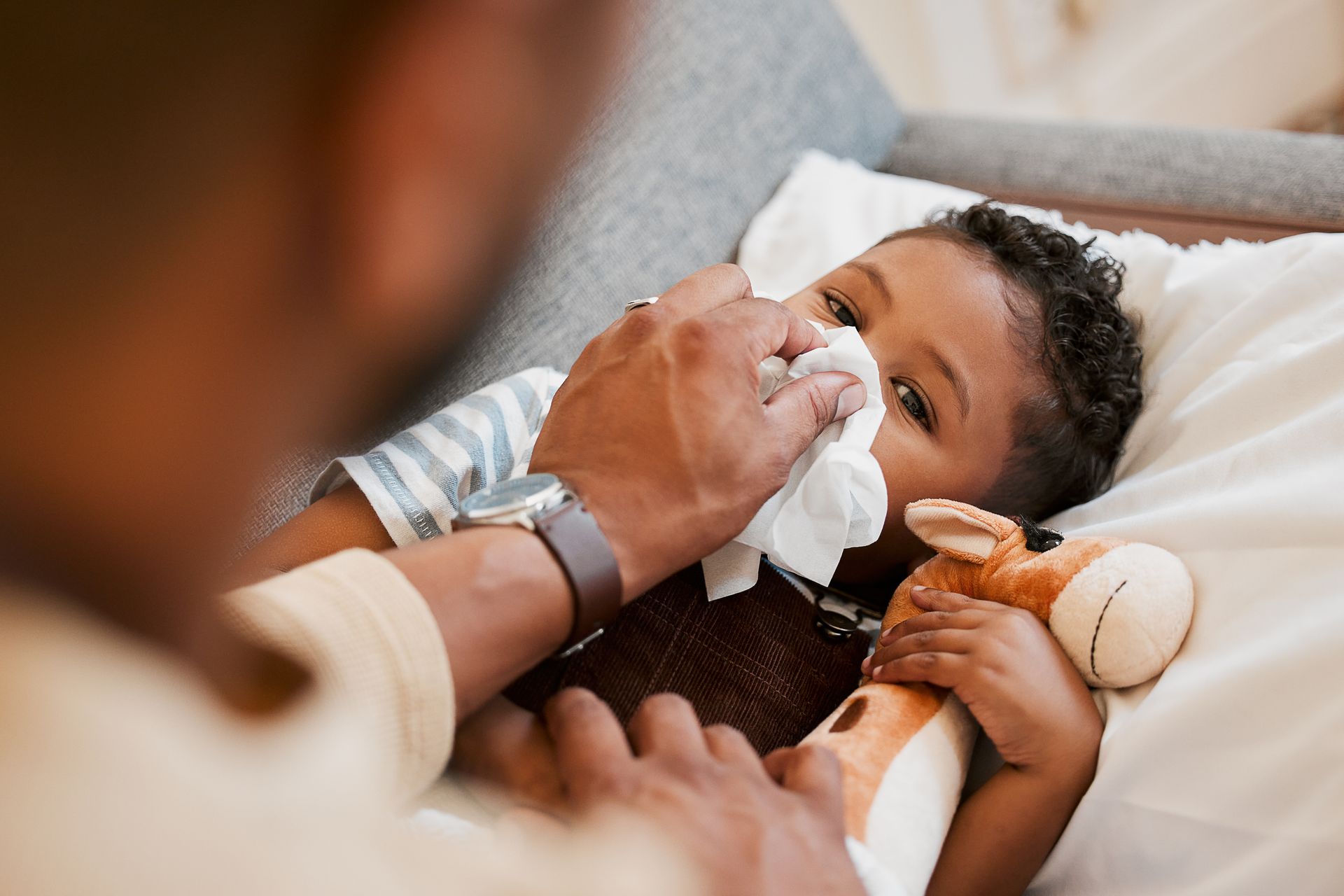 Sick little boy lying in bed while his parent wipes his nose with a tissue