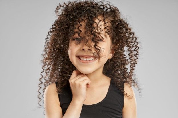 A little girl with curly hair is smiling and touching her face.