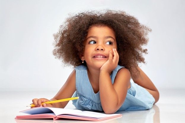 A little girl is laying on the floor with a book and a pencil.