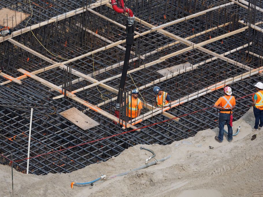 Construction workers on a rebar foundation grid at a building site, wearing orange safety vests and helmets.