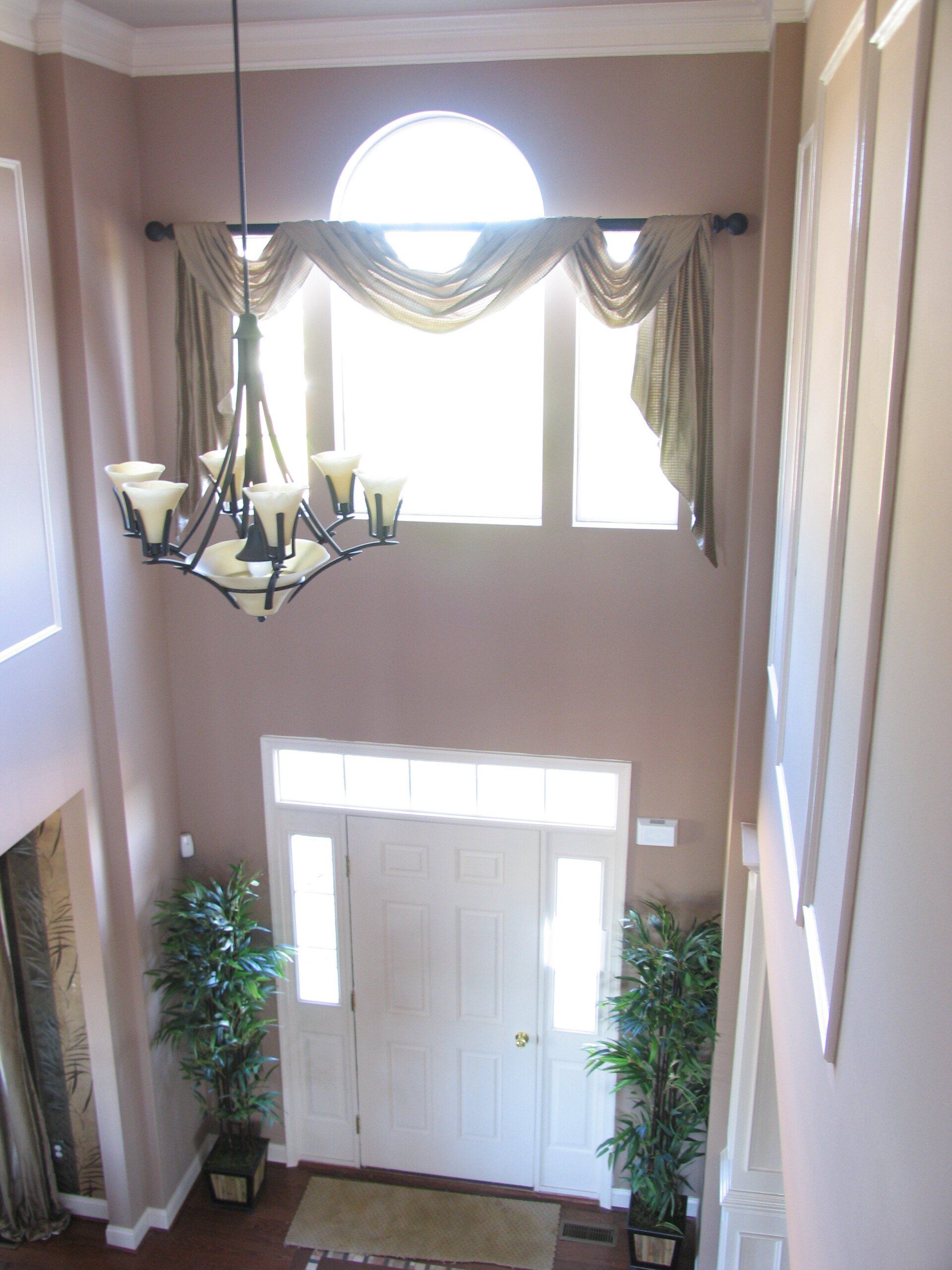 High-ceiling entryway with white door and windows, beige walls, chandelier, and draping curtains.