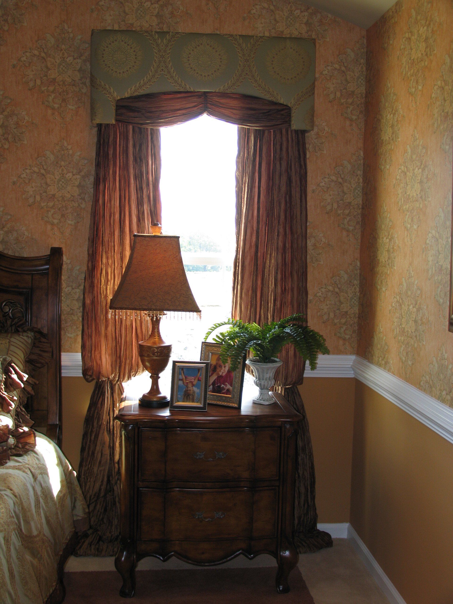 Nightstand with lamp and framed photos by a window with sheer, brown curtains and valance, against textured, orange walls.