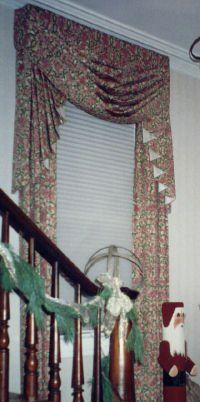 Window with floral drapery, blinds, and Christmas decor next to a staircase with garland and a Santa figure.