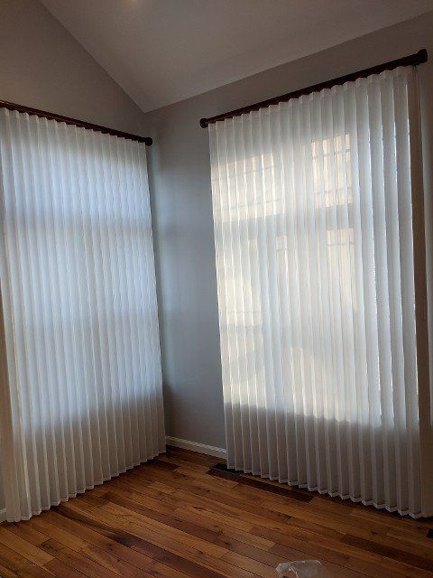 Two sheer white pleated curtains hang in a room with hardwood floors and gray walls.