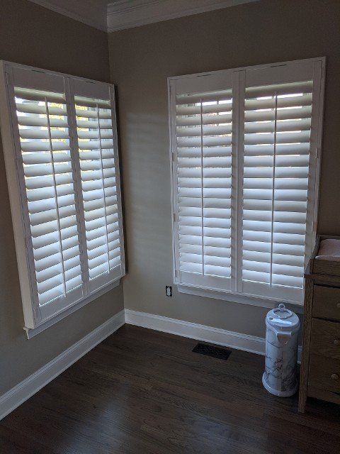Two white shuttered windows in a room with dark wood floor and light gray walls.