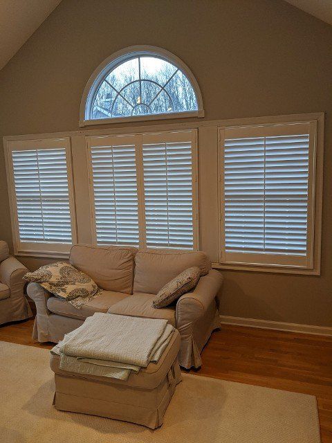 Living room with three windows, sofa, and ottoman; neutral tones, white shutters.