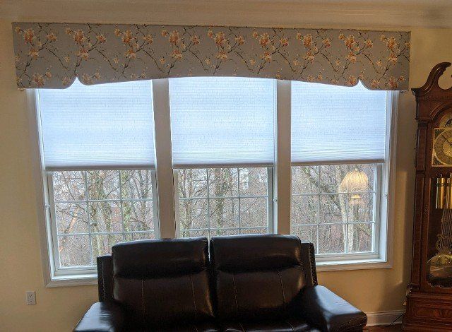 A living room with three windows, covered by blinds and a decorative valance, above a black couch.