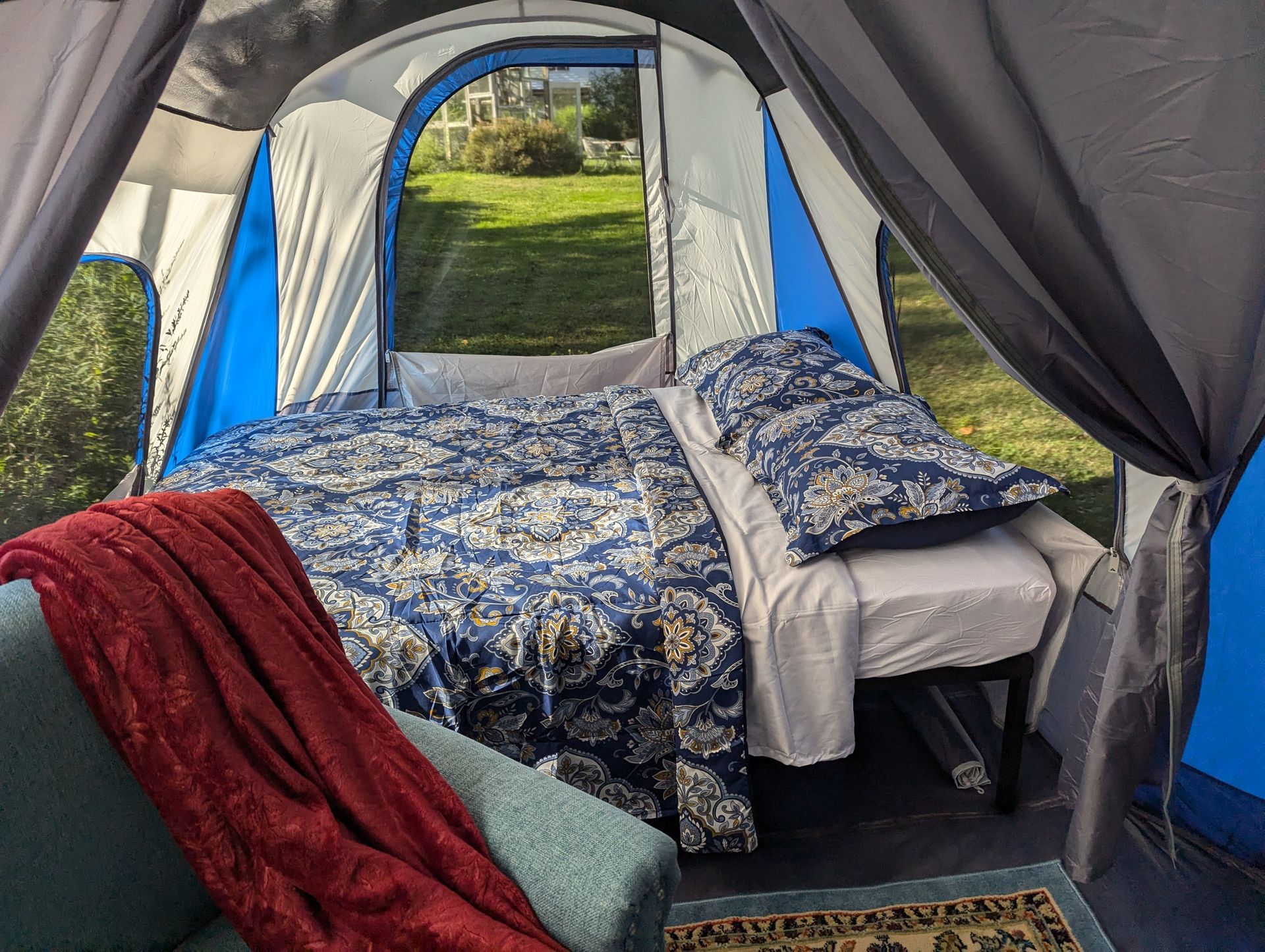 Inside a large tent, a bed with blue patterned bedding is set up. A red blanket and teal couch are nearby.