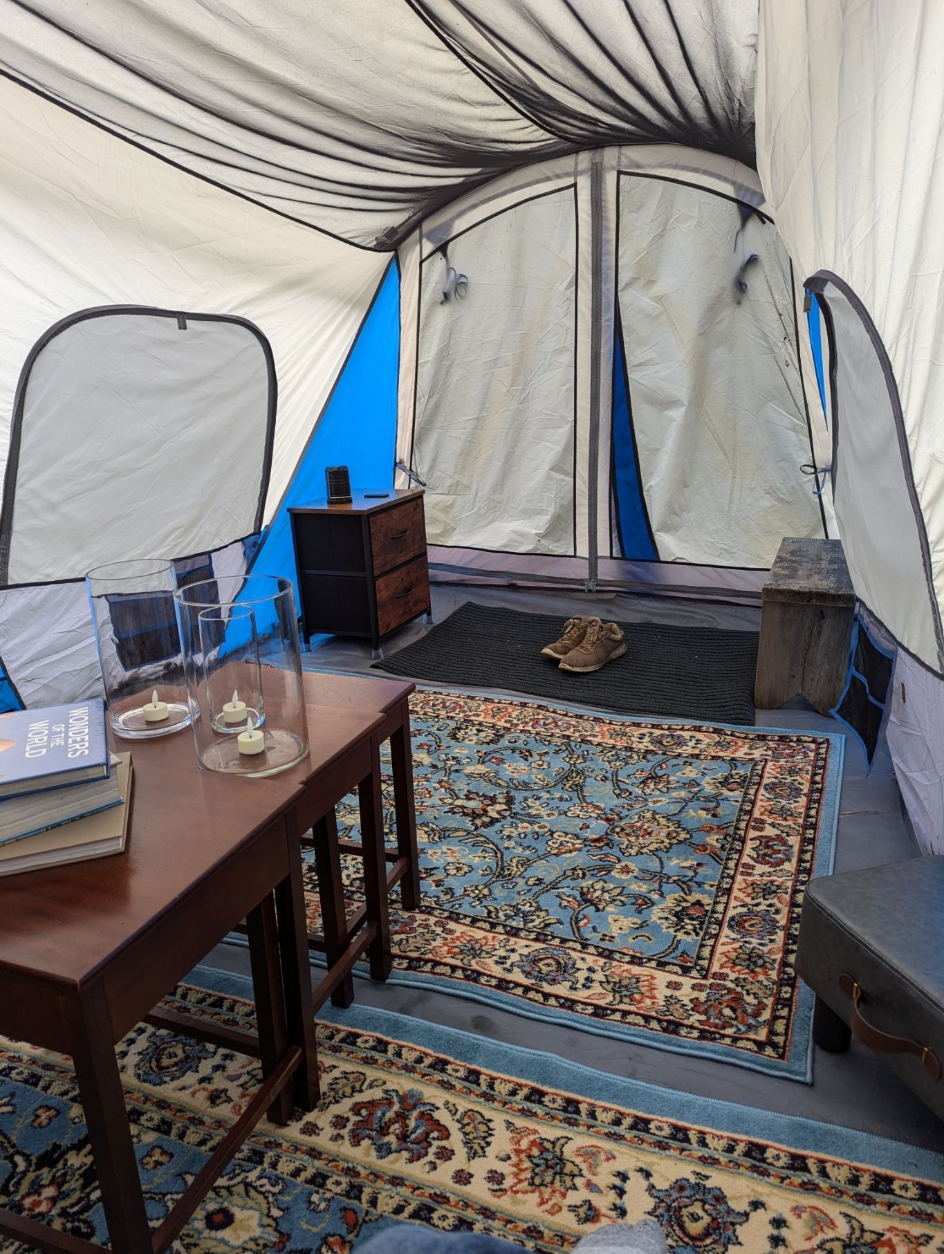 Inside a tent, a small table with books and candles, a rug, a dresser, and open tent flaps.