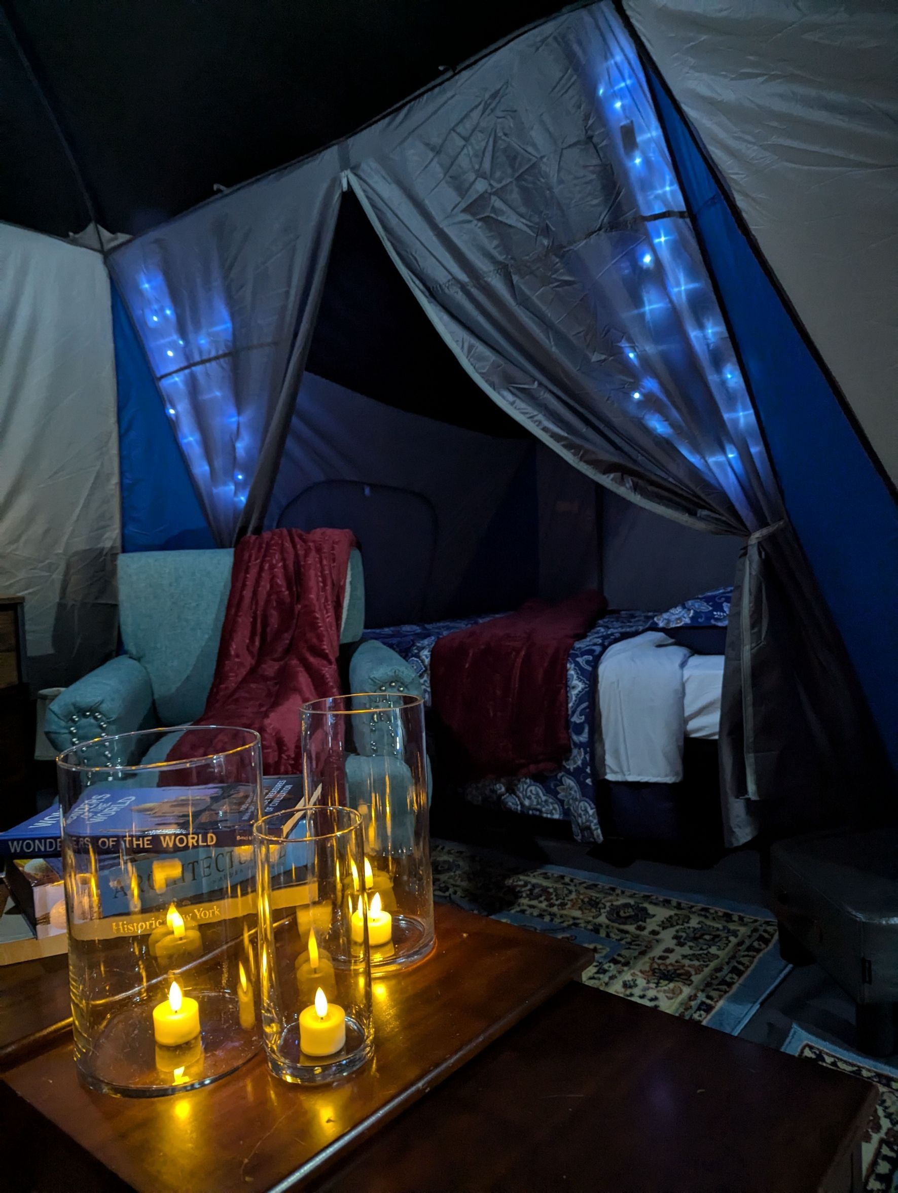 Cozy tent interior with bed, armchair, table with lit candles, and string lights.