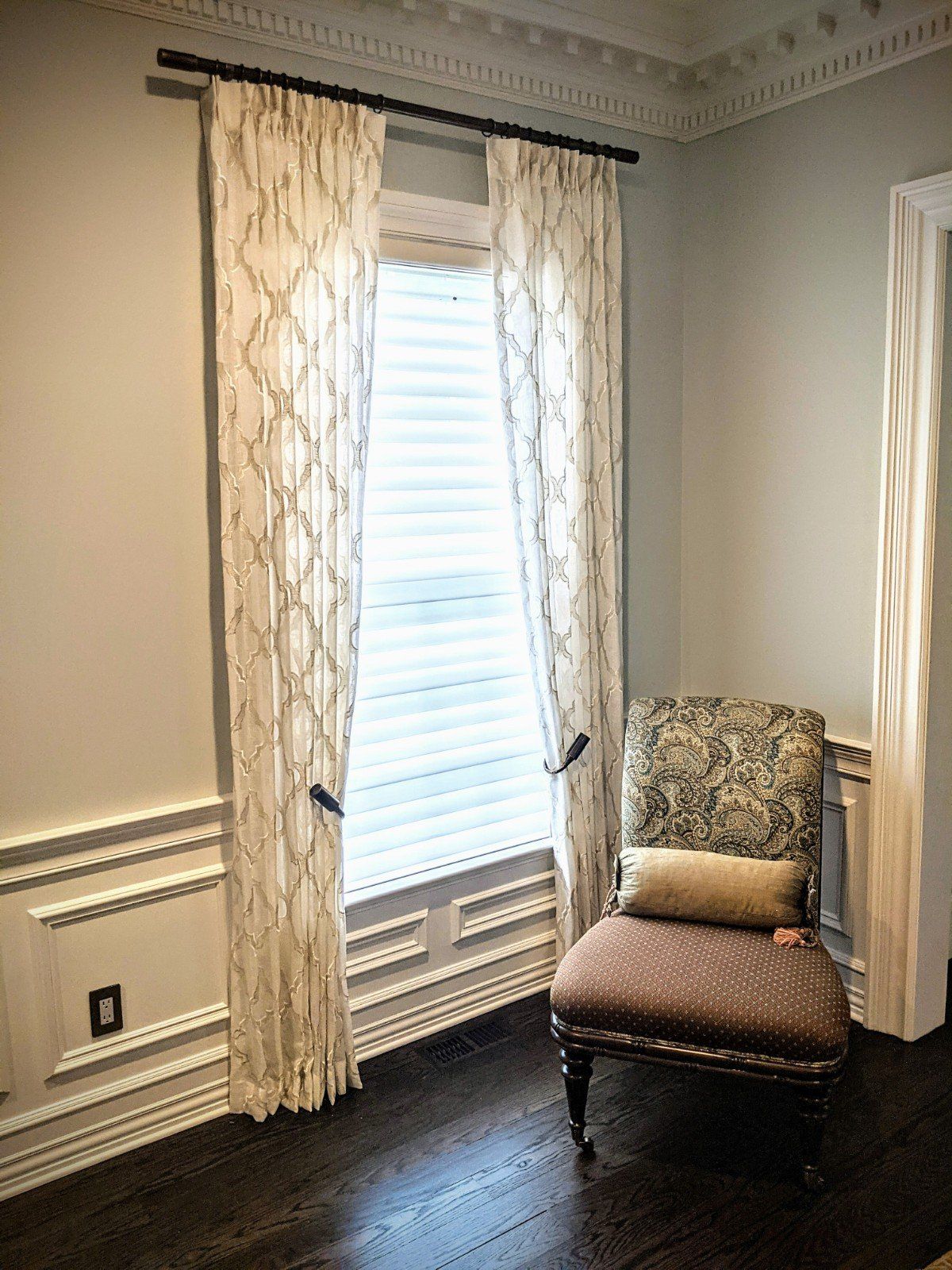 Window with patterned curtains, a chair, and decorative molding in a room with dark wood floors.
