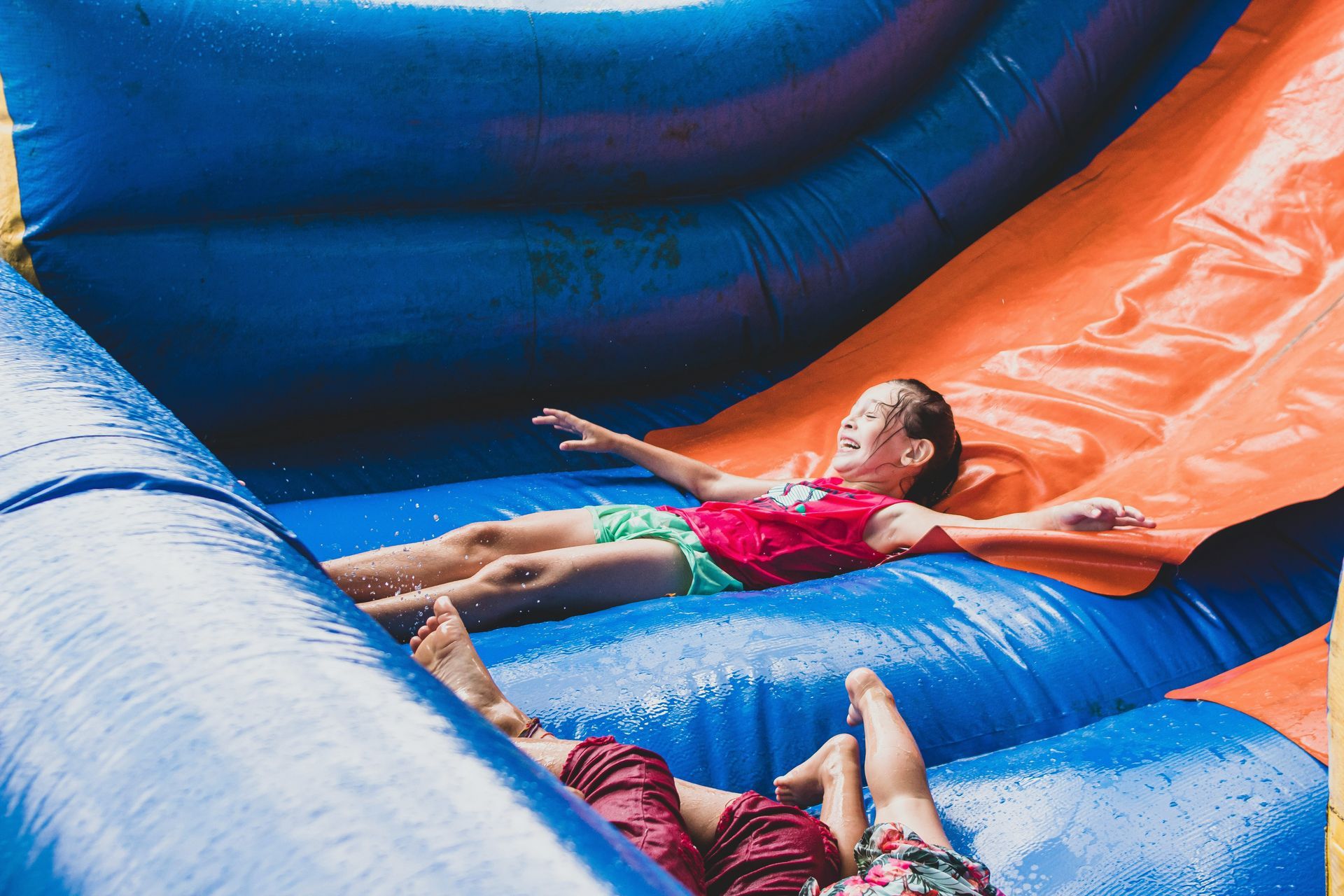 Children sliding on a blue and orange inflatable water slide, arms outstretched and smiling