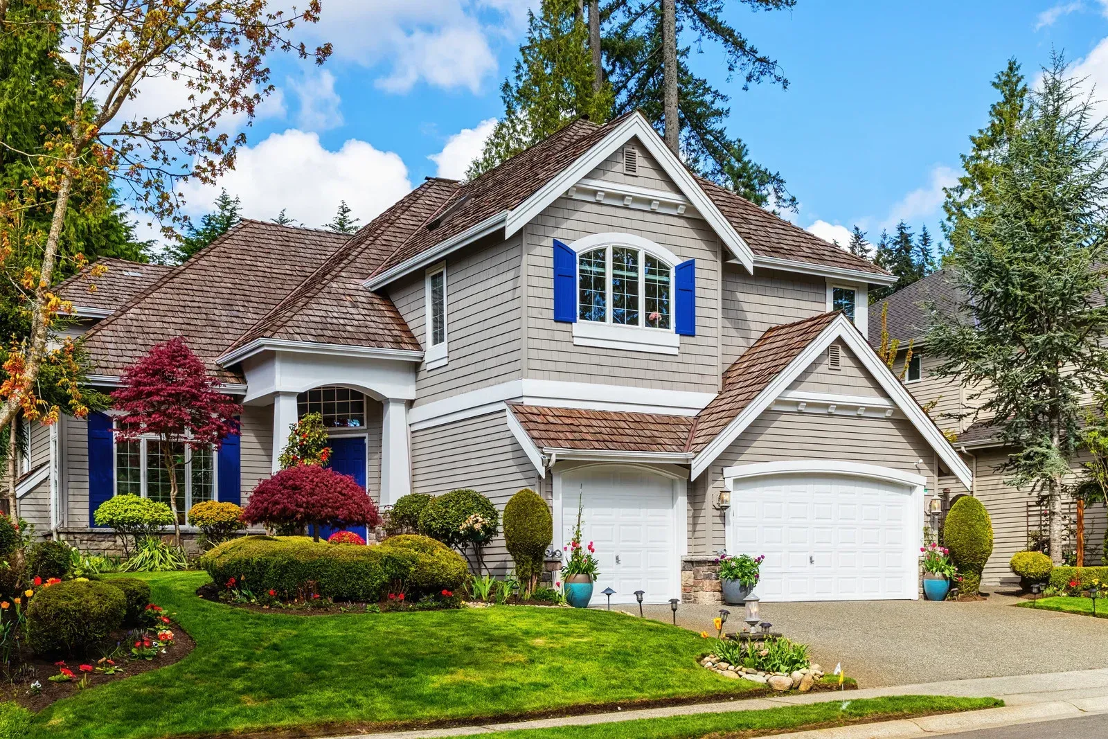Two-story house with gray siding, white garage doors, blue shutters, and well-landscaped yard under a sunny sky.