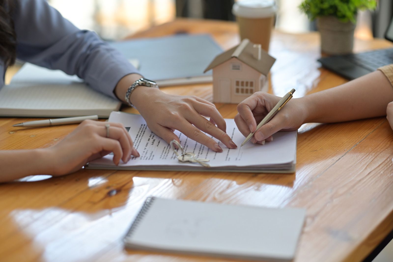 Two people signing a contract at a wooden table with a house model, a coffee cup, and a laptop.