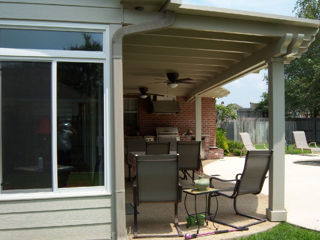 Covered patio with dining set, overlooking a pool. Brick wall, tan posts and roof, sunny outdoor setting.