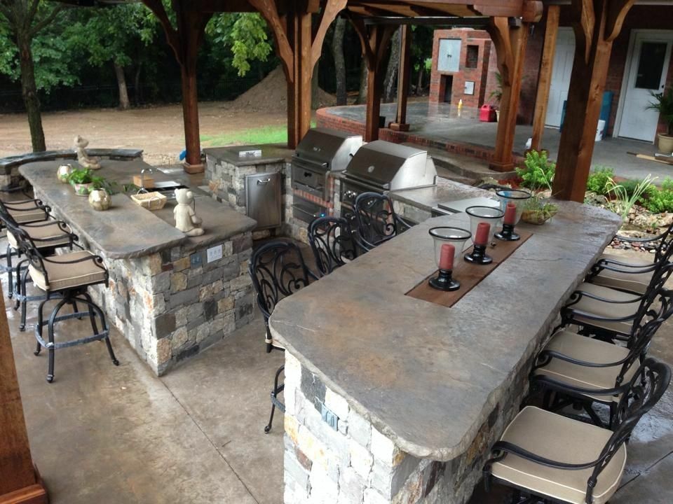 Outdoor kitchen with stone counters, built-in grills, and bar seating under a wooden pergola.