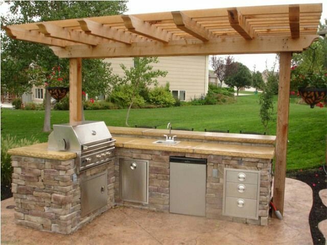 Outdoor kitchen with grill, sink, and storage under a wooden pergola on a patio, overlooking a green lawn.