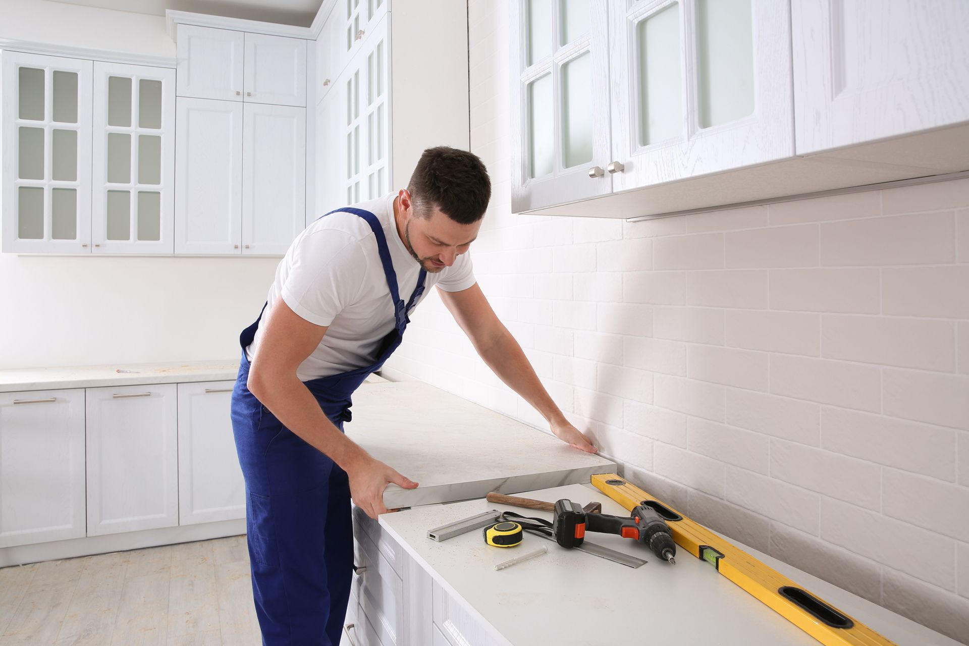 Man in blue overalls installing countertop in a white kitchen with tools.