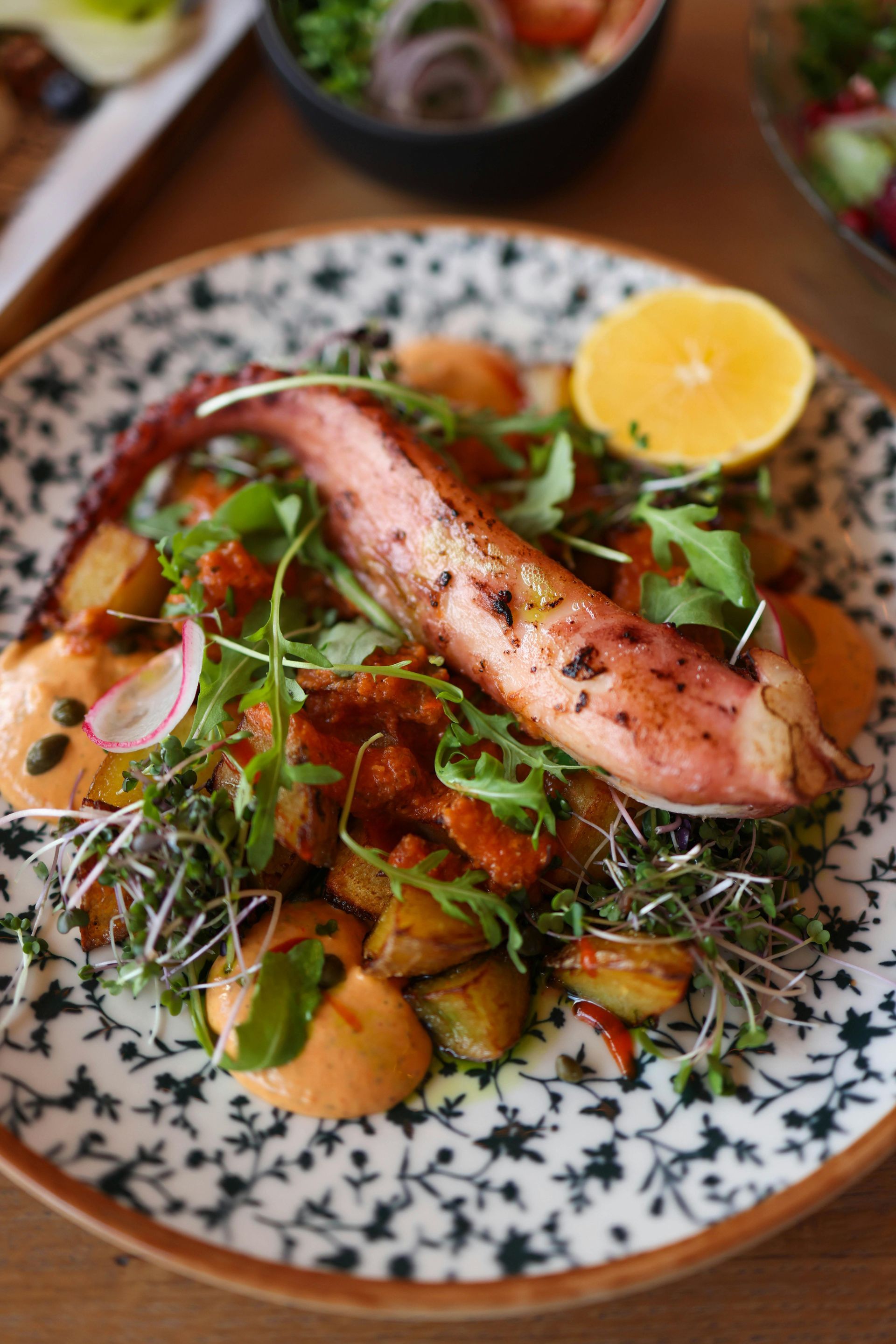 Grilled octopus tentacle on a plate with roasted potatoes, greens, sauce, and a lemon wedge.