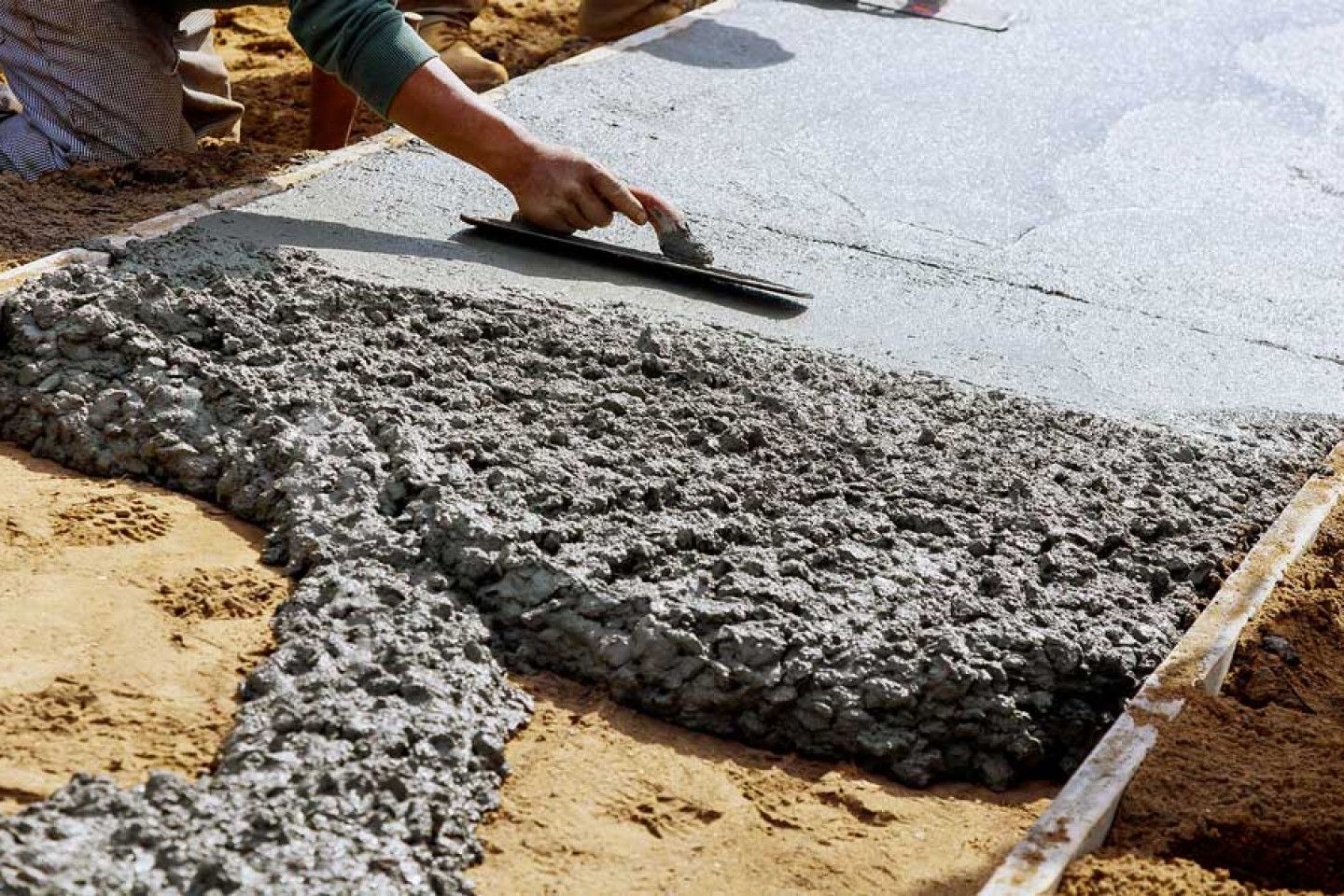 Person smoothing wet concrete with a trowel, creating a flat surface within a wooden frame on a sandy ground.