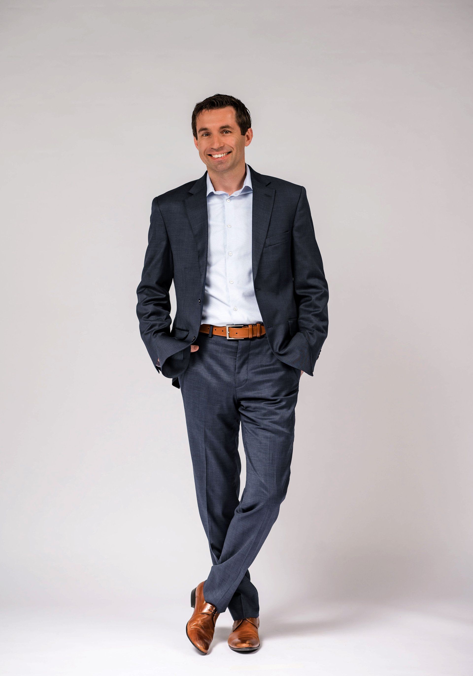 Man in navy suit and white shirt, standing with hands in pockets against a plain gray background.