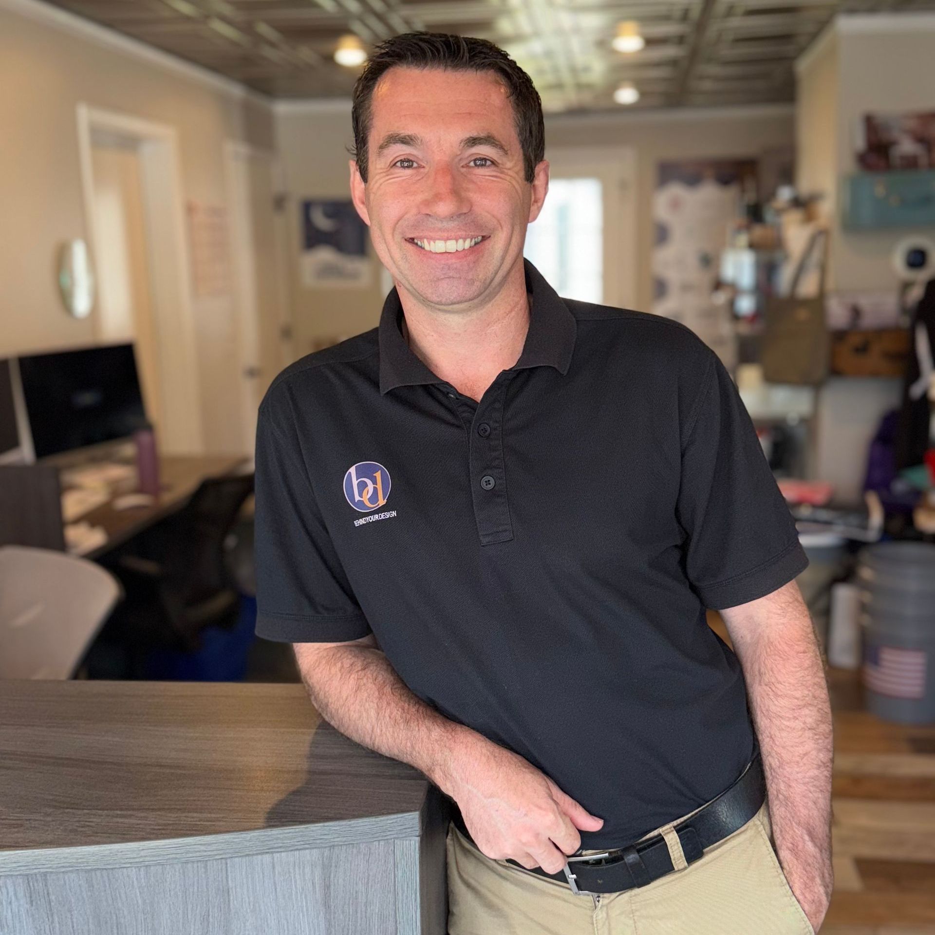 Smiling man in a black polo shirt leaning on a counter in a bright office lobby