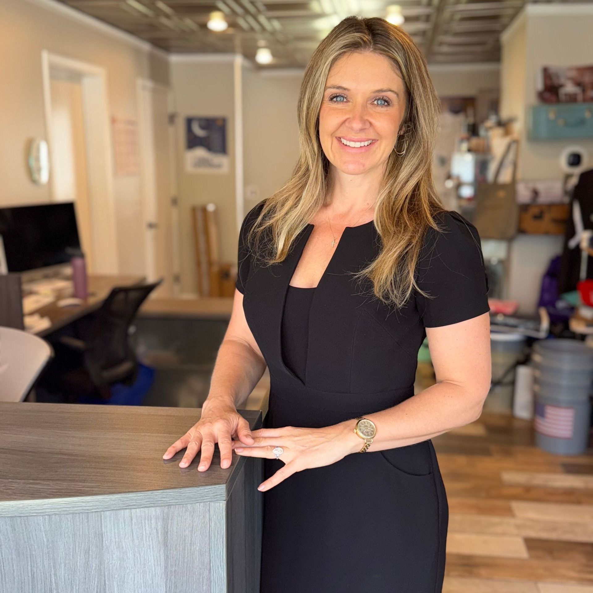 Woman standing at a salon reception desk, smiling in a black dress.