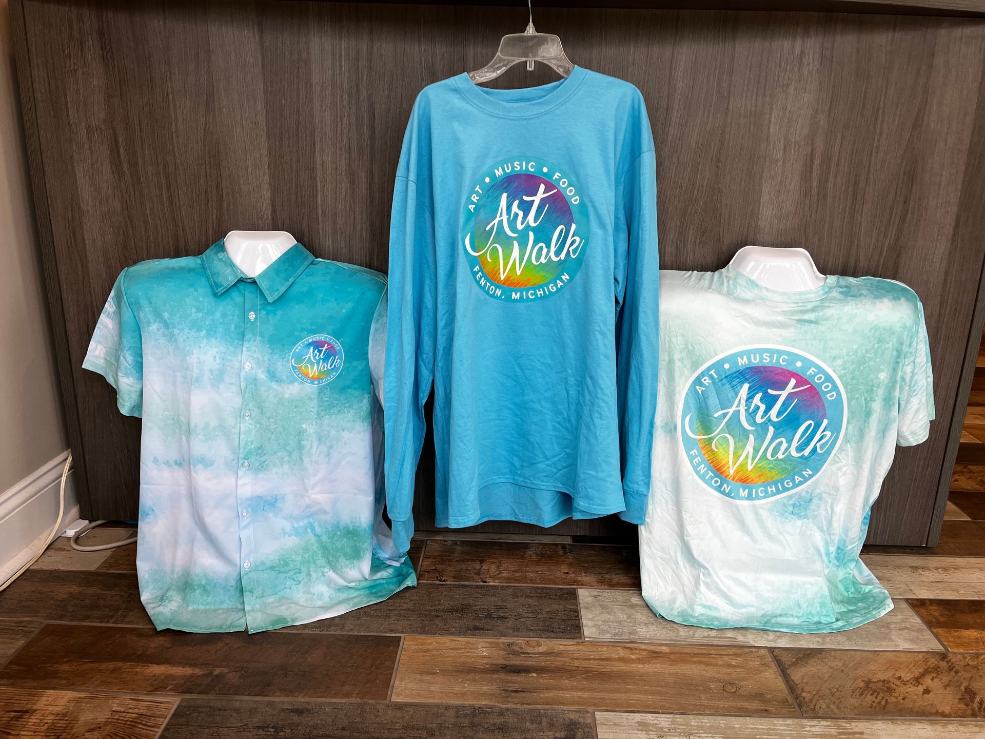 Three tie-dye Art Walk shirts displayed on mannequins and hangers against a wood wall