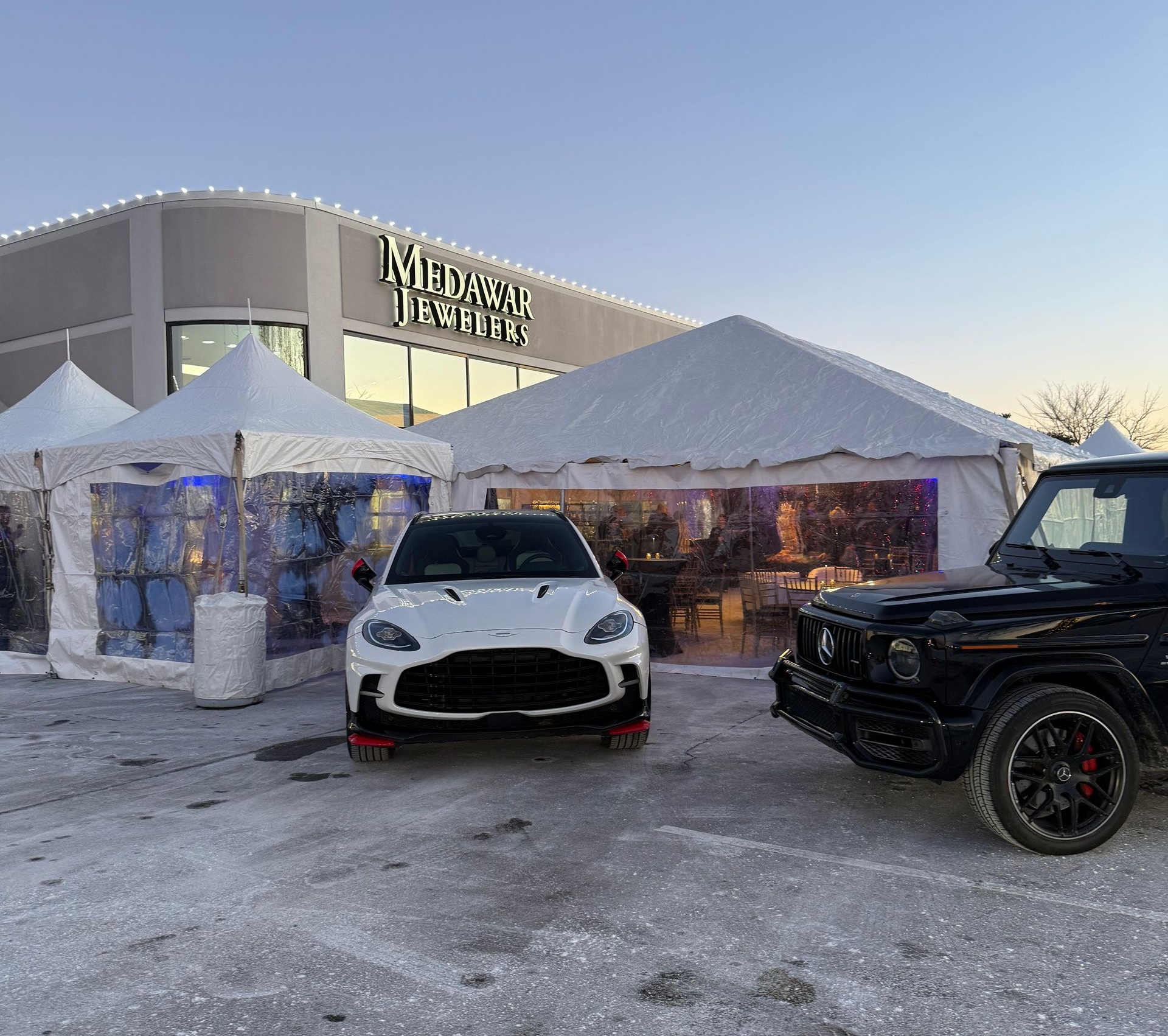 White sports car and black SUV outside a tented winter event at dusk