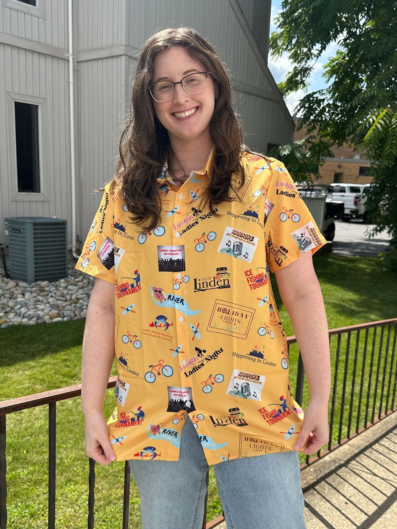 Smiling person in a yellow patterned shirt standing outdoors by a building and railing.