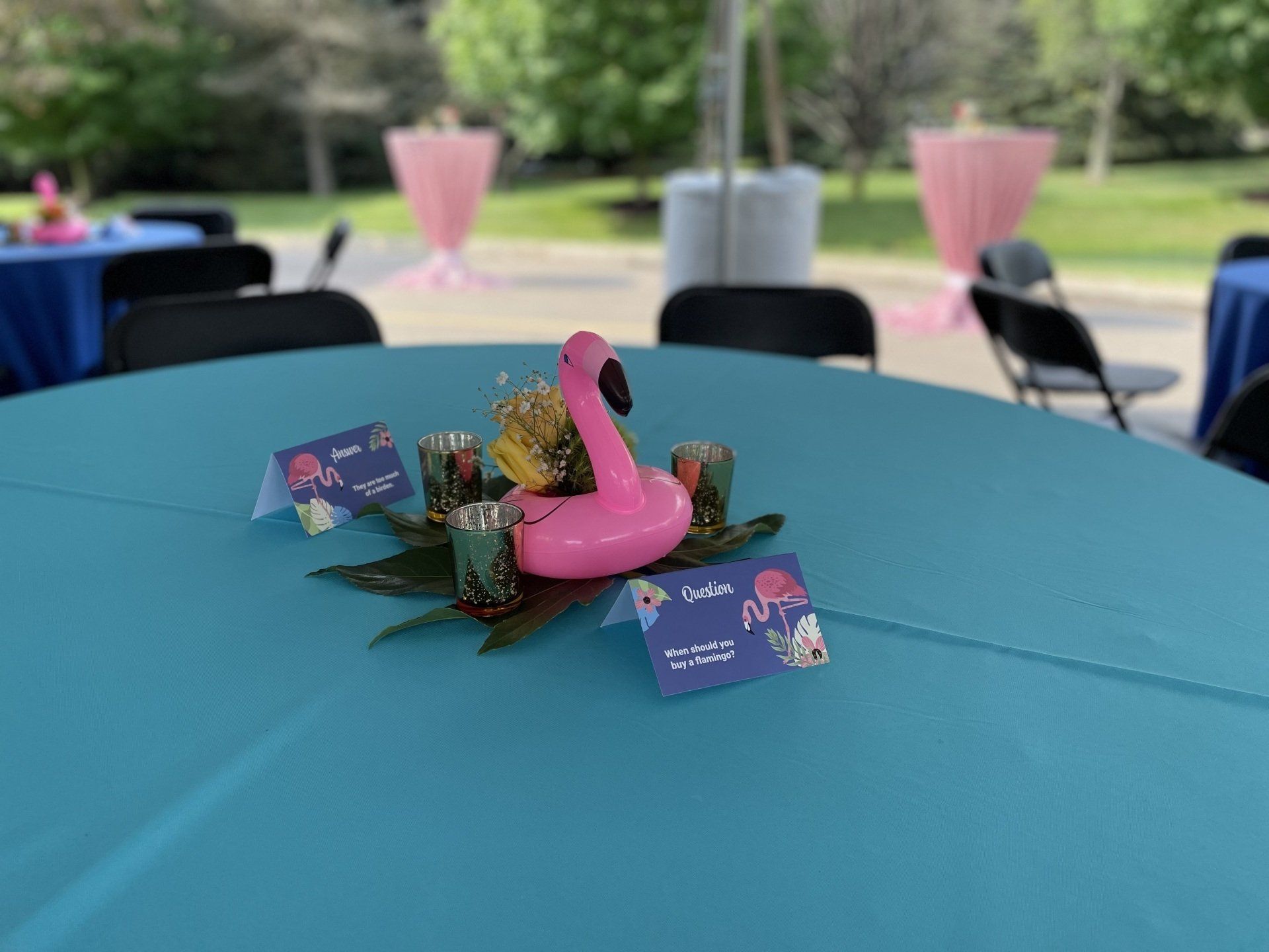 Pink flamingo centerpiece on a teal table at an outdoor event with pink napkins and cards