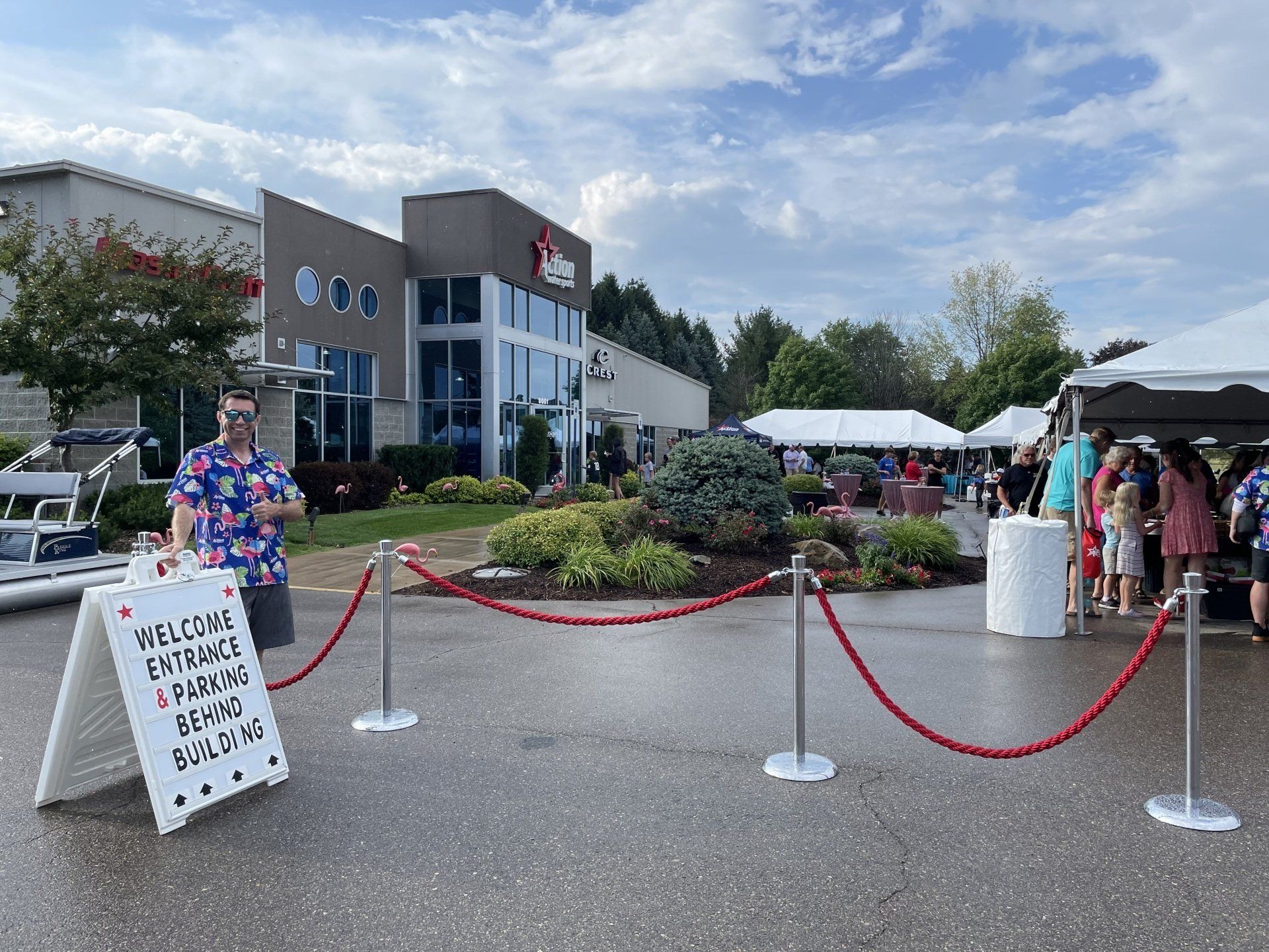 Outdoor market entrance with red rope barriers, white tents, and a sign on a wet parking lot.