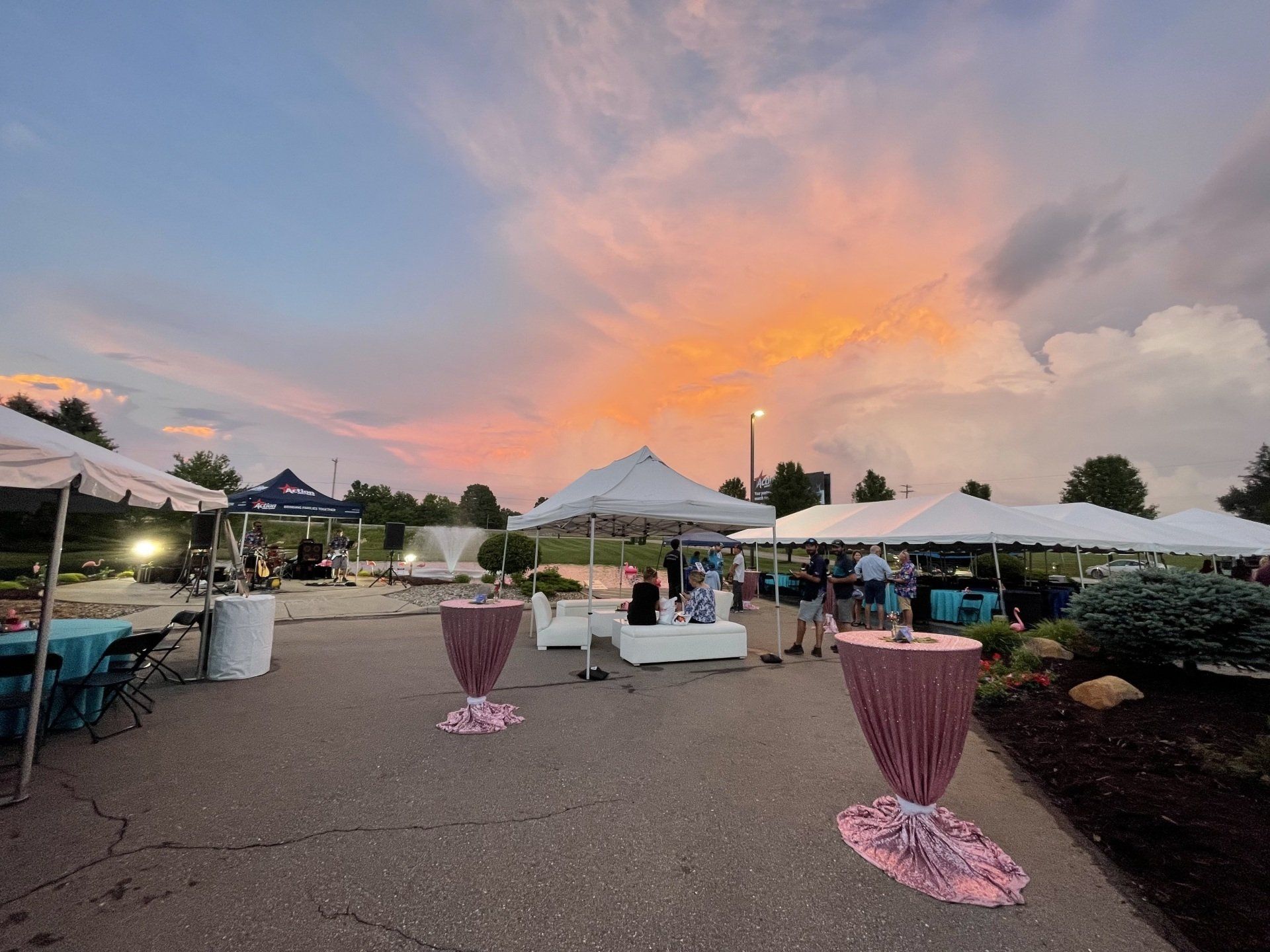 Outdoor lounge area at sunset with white tents, tables, and pink-draped stands under a colorful sky