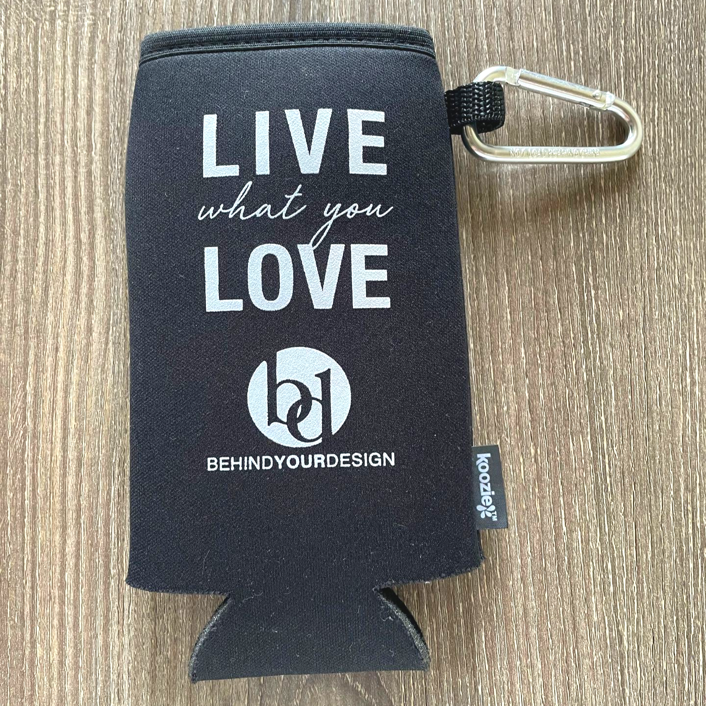 Black can koozie with “LIVE what you LOVE” text and a metal clip on a wooden surface