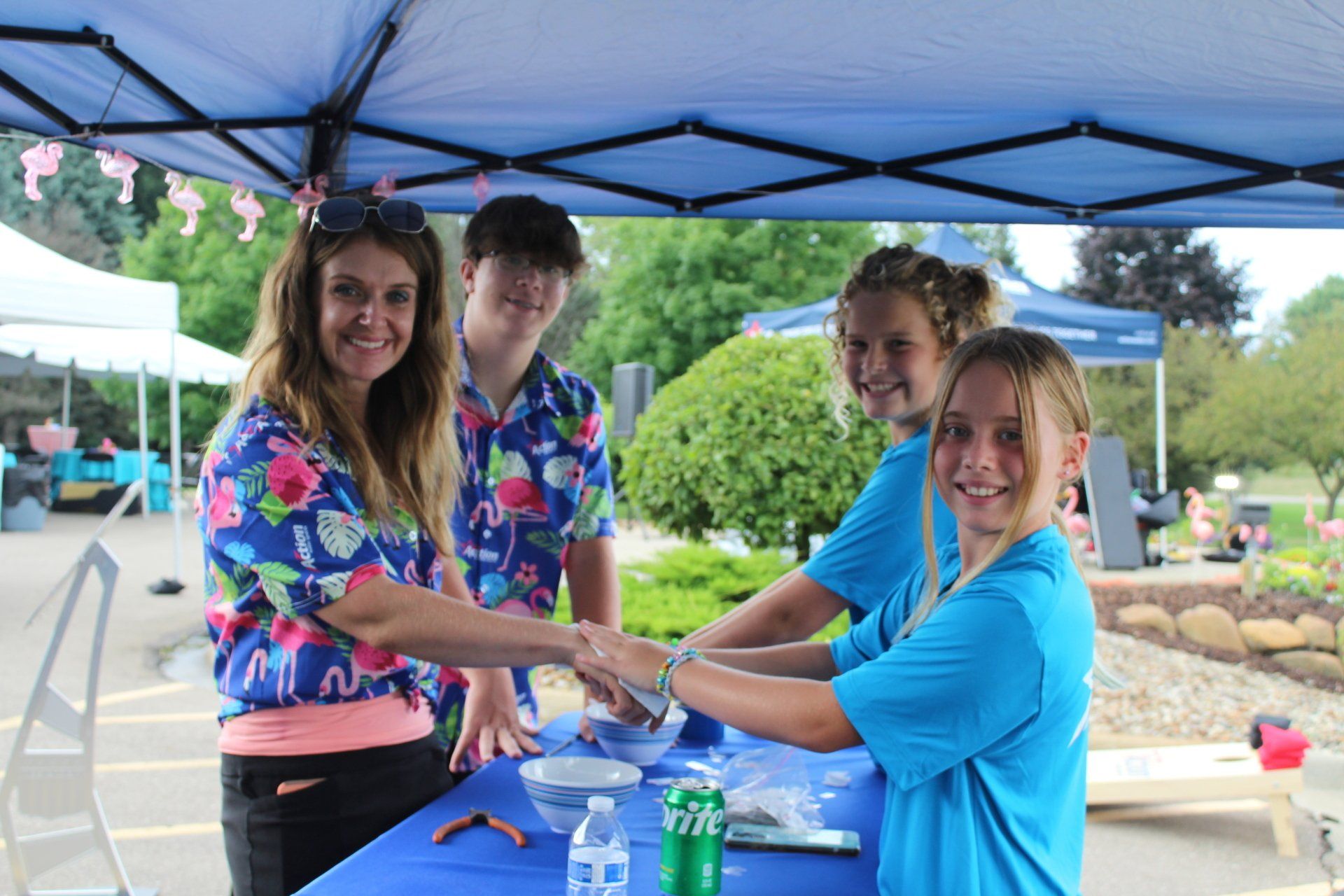 Four people in colorful shirts at a blue-canopy table, stacking hands and smiling outdoors.