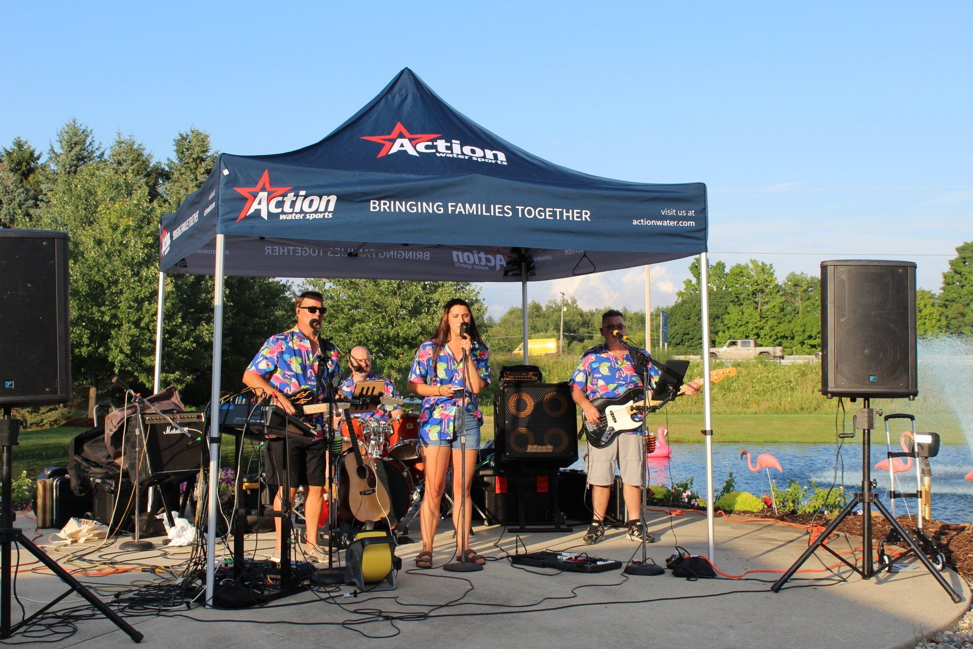 Three musicians performing on an outdoor stage under a blue canopy, with speakers and drums nearby.