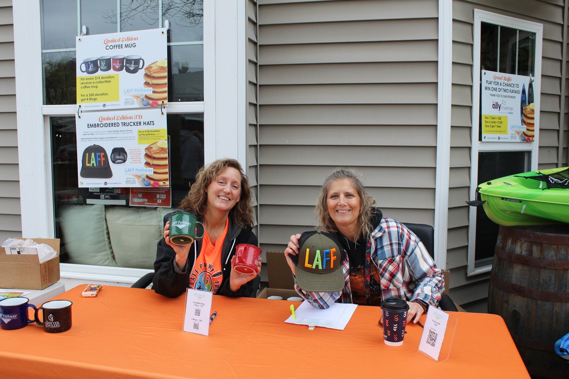 Two women seated at an orange table at a community event booth with posters in the background