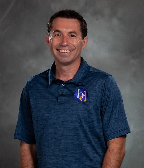 Smiling man in a blue polo shirt with a logo, standing against a gray studio backdrop