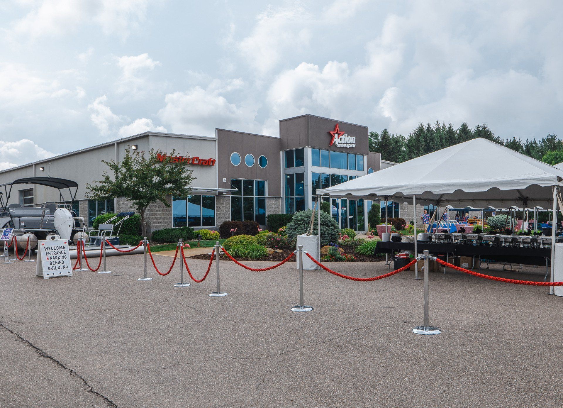 Outdoor event area in front of a building, with red rope barriers, tents, and parked cars under cloudy skies