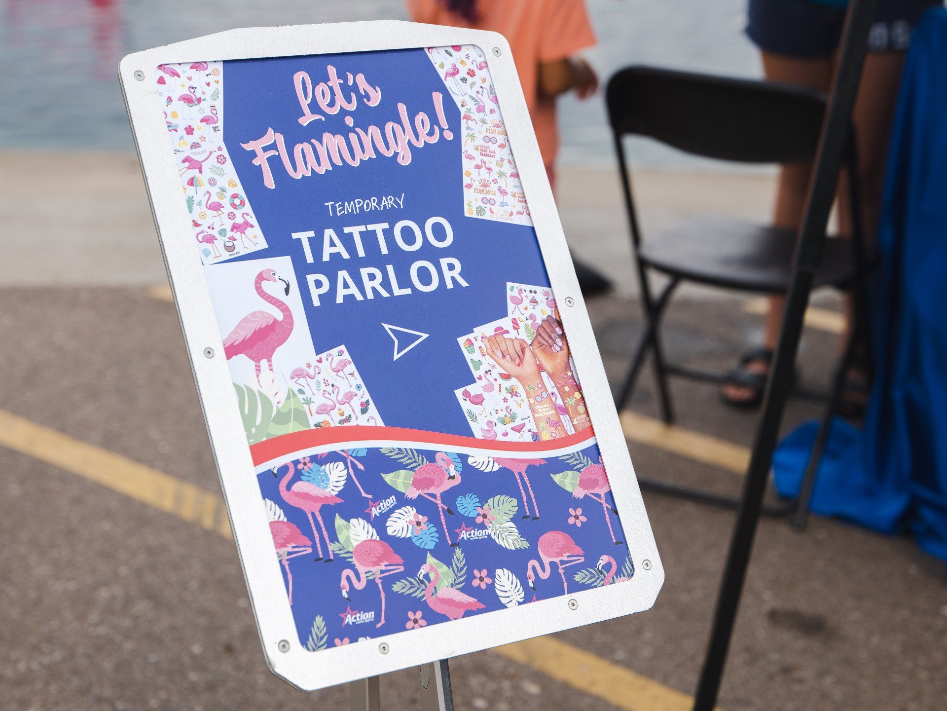 Handmade sign for “The Tattoo Parlor” with a flamingo design on a sidewalk near chairs