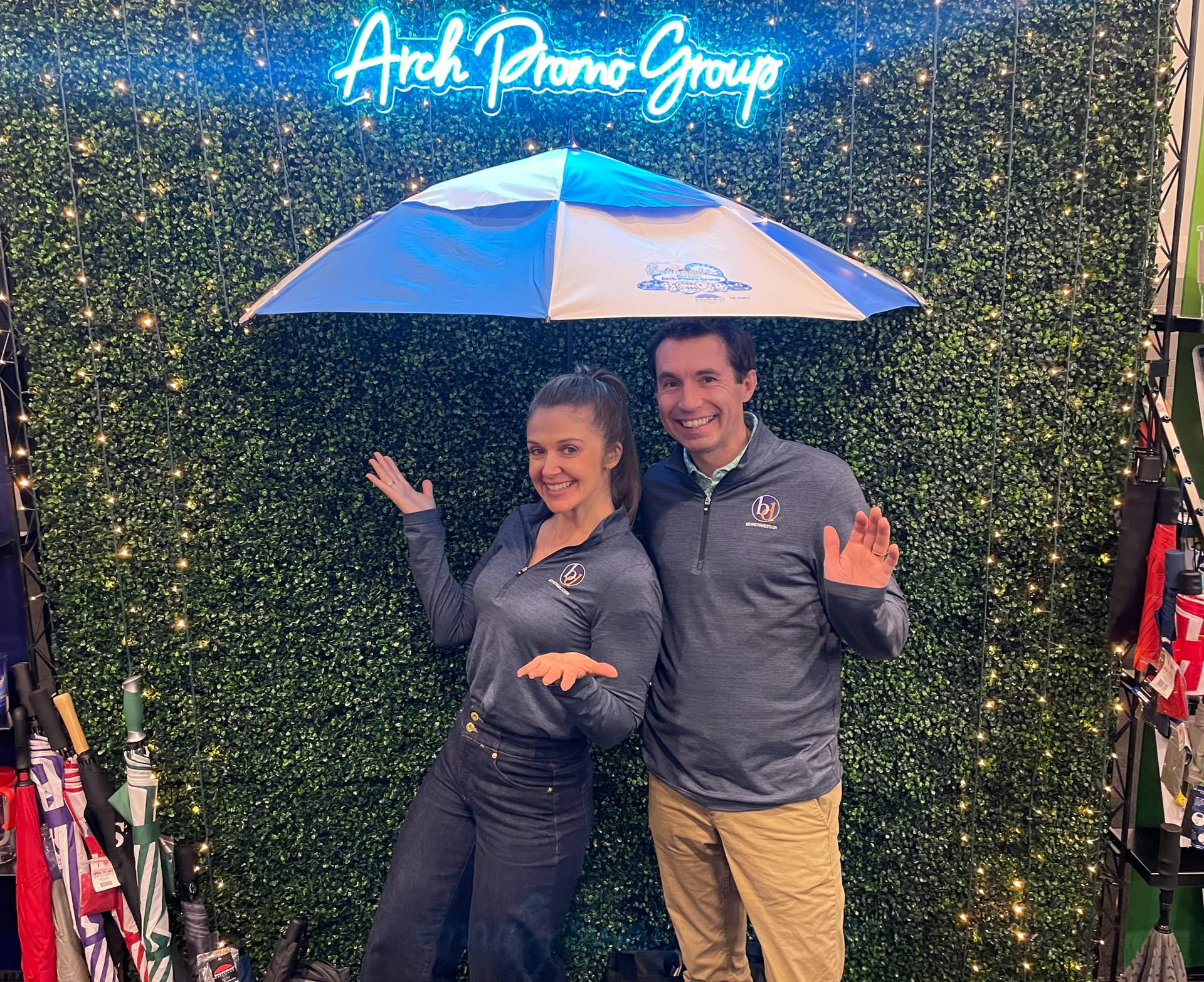 Two people posing under an umbrella in front of a green hedge wall and neon sign