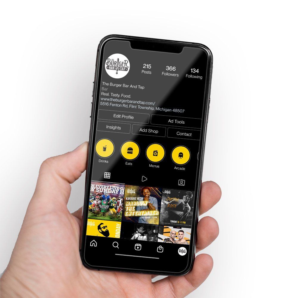Hand holding a smartphone showing a dark-themed social media profile page with yellow icons and photo grid.