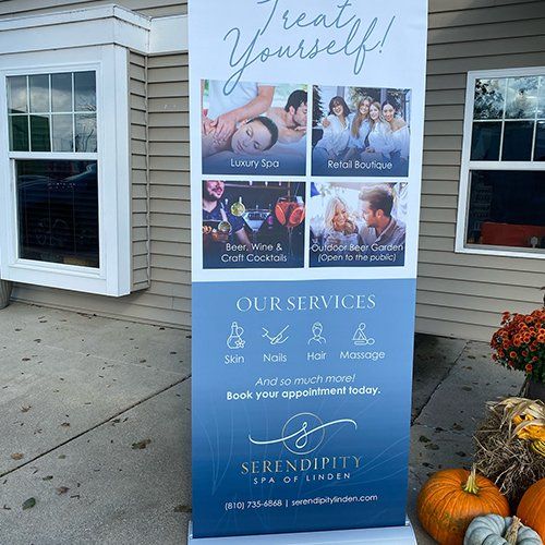 Blue spa services sign on a sidewalk beside a building, with wellness photos and pumpkins nearby.